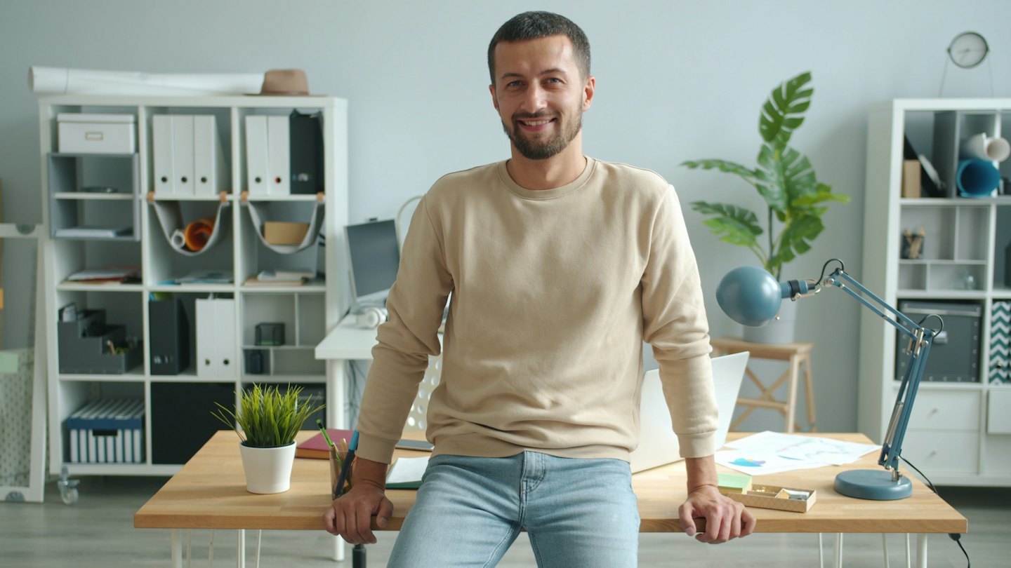 Uomo con maglione beige appoggiato a una scrivania d'ufficio.